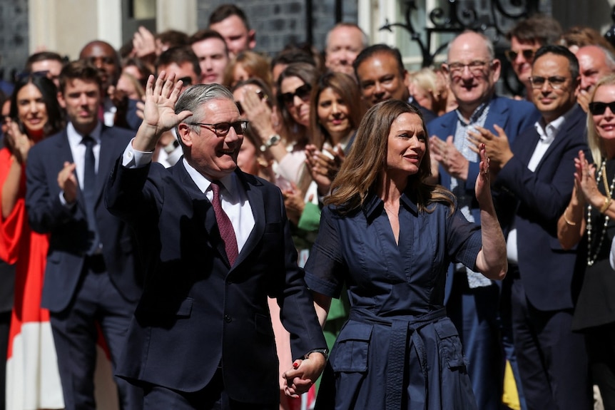 A man and a woman hold hands, wave and smile. A large crowd clapping is visible behind them.
