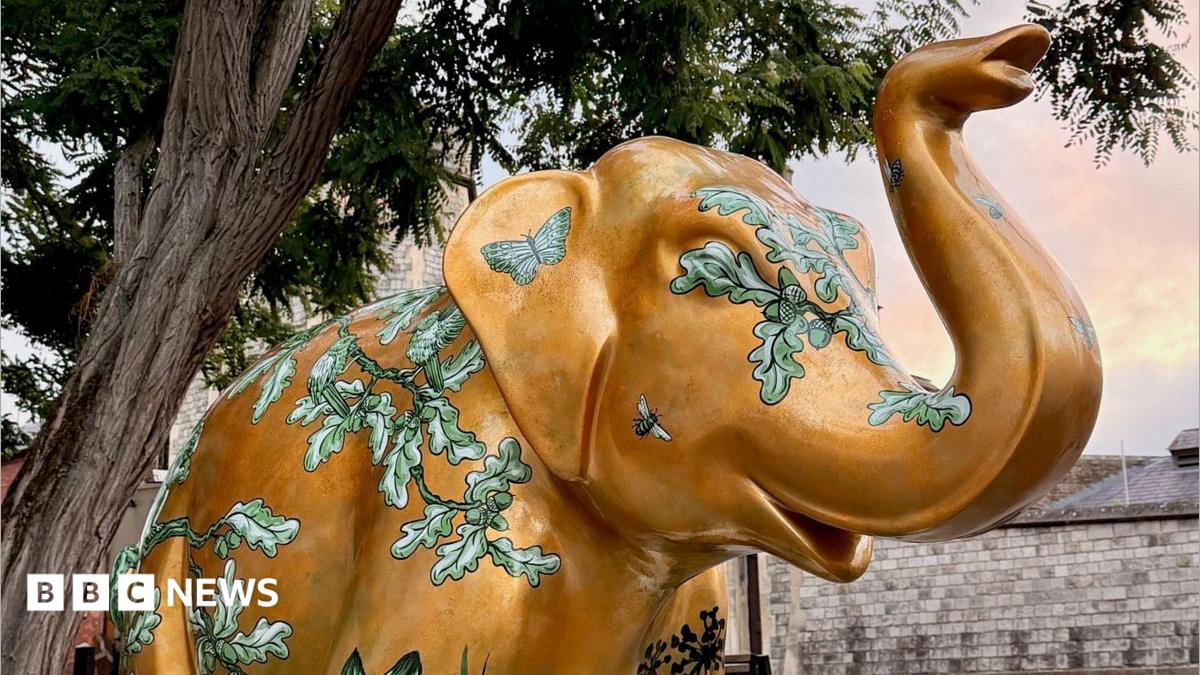 An elephant sculpture is pictured next to a tree. It is gold in colour with green leaves and foliage painted around its body.
