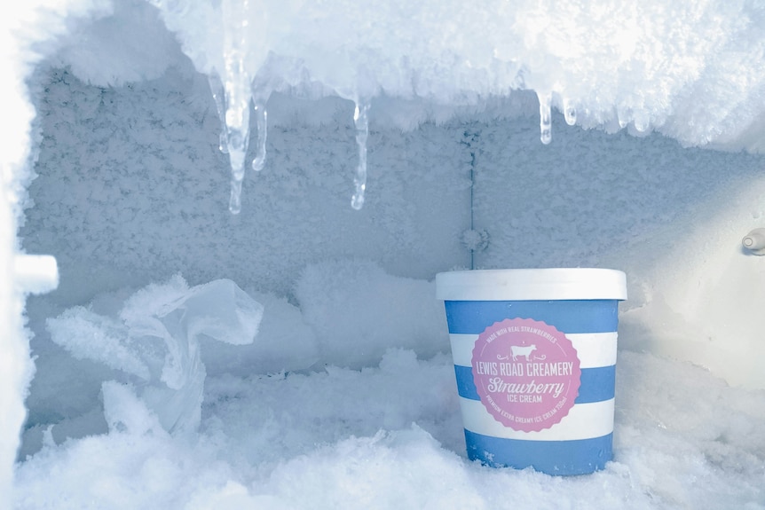 A pint of ice cream inside a freezer coated in thick ice, with icicles extending from the roof.