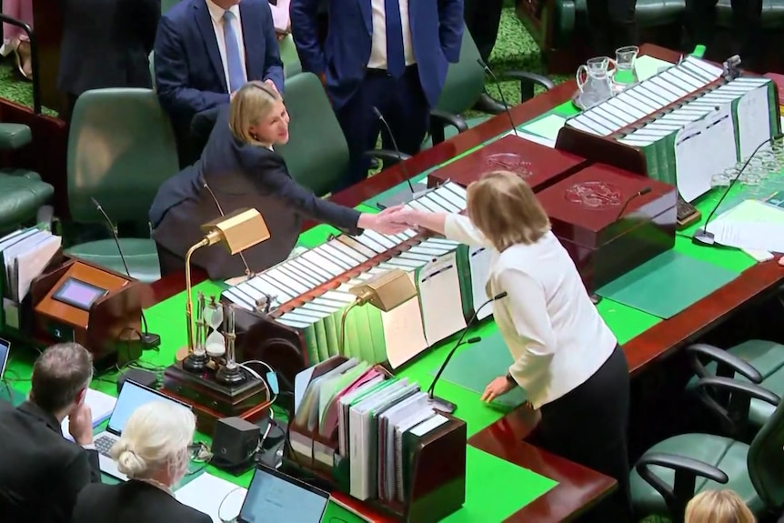 A blonde woman in a suit and a brown haired woman in a cream jacket lean across a large table covered in books and shake hands.