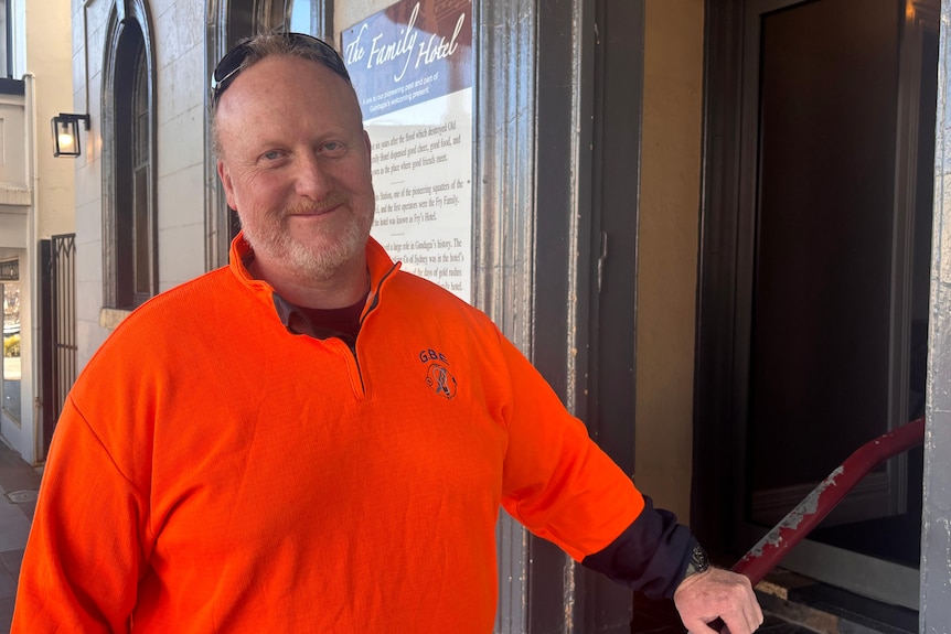 Man in a fluro orange jumper and sunglasses on his head poses in front of the steps of a pub