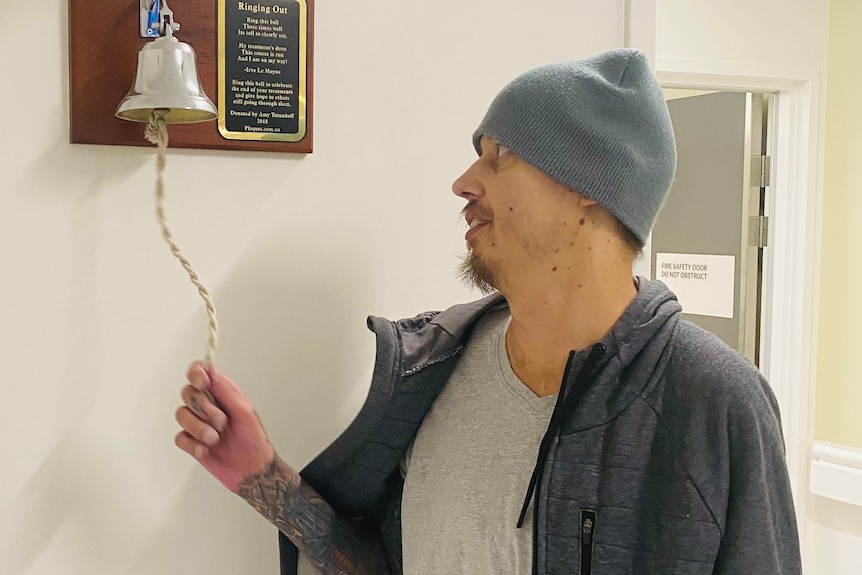 A man wearing a woollen beanie ringing a bell attached to a wall.