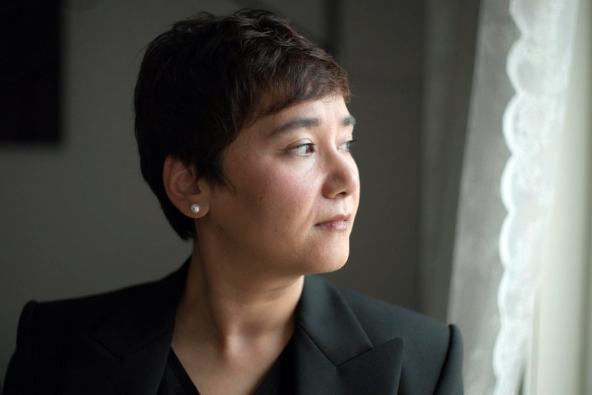 An Asian woman with short dark hair looking out of a window. She has a serious expression.