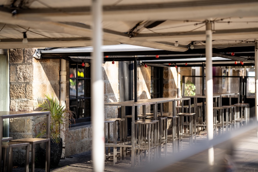 High tables and stools under umbrellas out the front of a sandstone building with plants and red fairy lights.