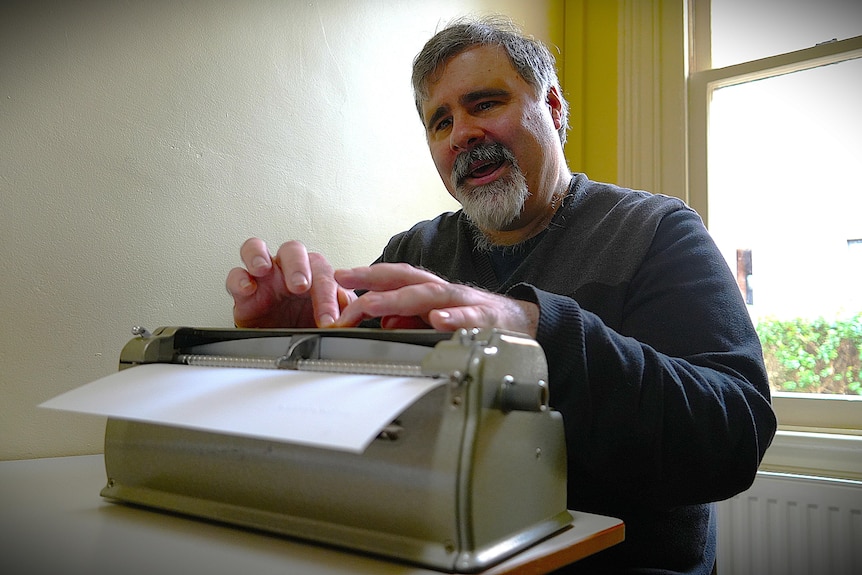 Man with grey hair and facial hair typing on a braille music writer