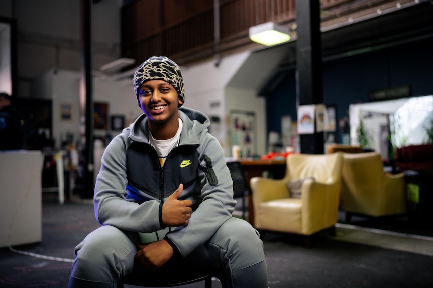 Befiker, wearing a grey tracksuit and a cheetah print beanie, sits on a chair smiling with a thumbs up, purple light reflects