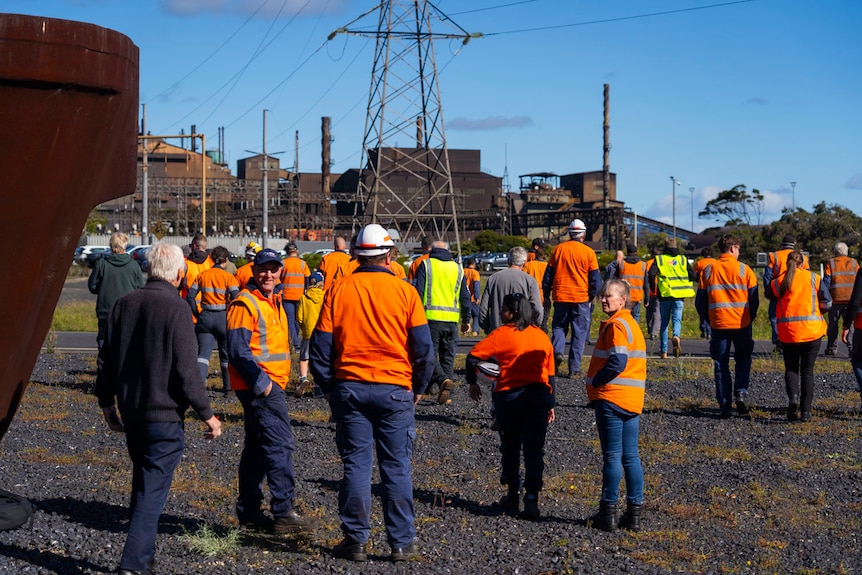 men and women in orange high vis outside a blue logo and a rusted sculpture