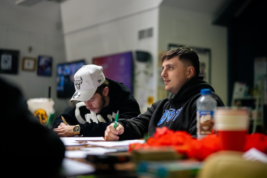 Two young men at a table writing with pen and paper, one wears a black jacket and looks up, chatting to someone out of frame.