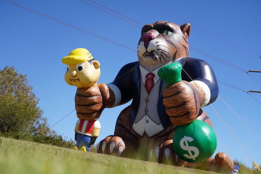 A large blow up cat in a suit, holding a bucket of cash in one hand and a worker in the other. The cat is smoking an cigar.