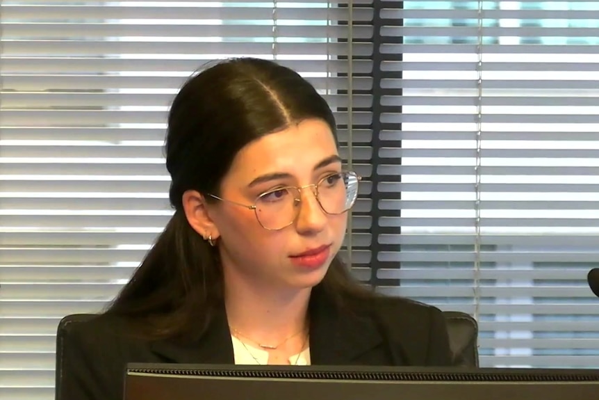 A woman with long dark hair pulled back in a ponytail in a black jacket with clear rimmed glasses sits behind two microphones.