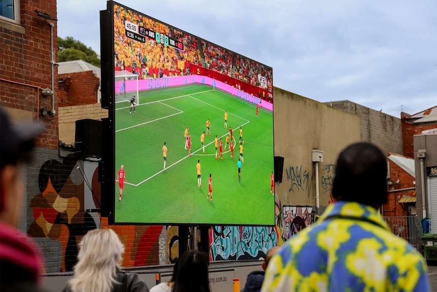 A large screen, showing a soccer pitch with players in the background, people can be seen in the foreground looking up at it.