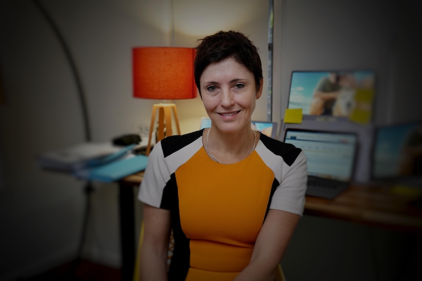 A white woman with a black pixie cut sitting at her office desk wearing orange, white, black top looking right of frame.