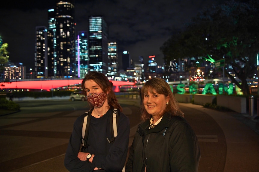 A woman smiles and another younger woman with a face mask on crosses her arms against a city backdrop at night