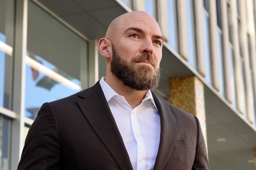 A bald man wearing a navy suit jacket stands outdoors looking serious.