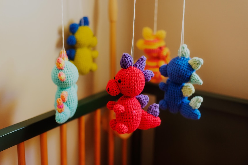 A mobile featuring colourful knitted dinosaurs hangs over a baby's cot.