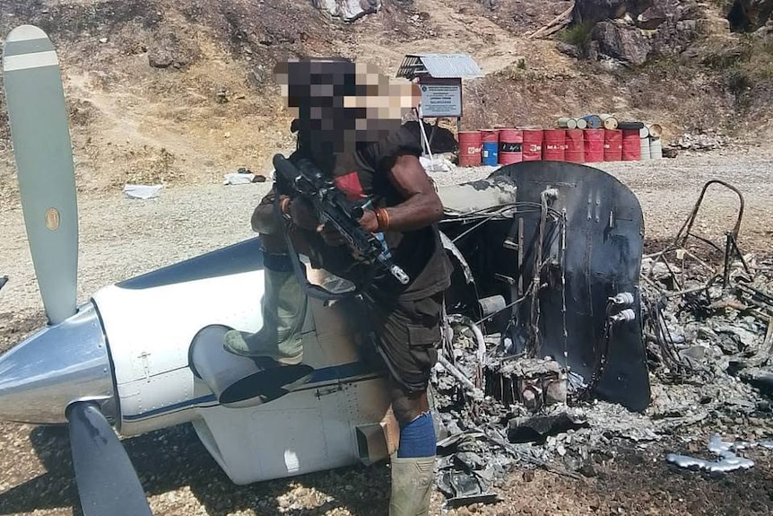 A man with a blurred face holds a gun while sitting on a burnt plane with red barrels behind him in the distance