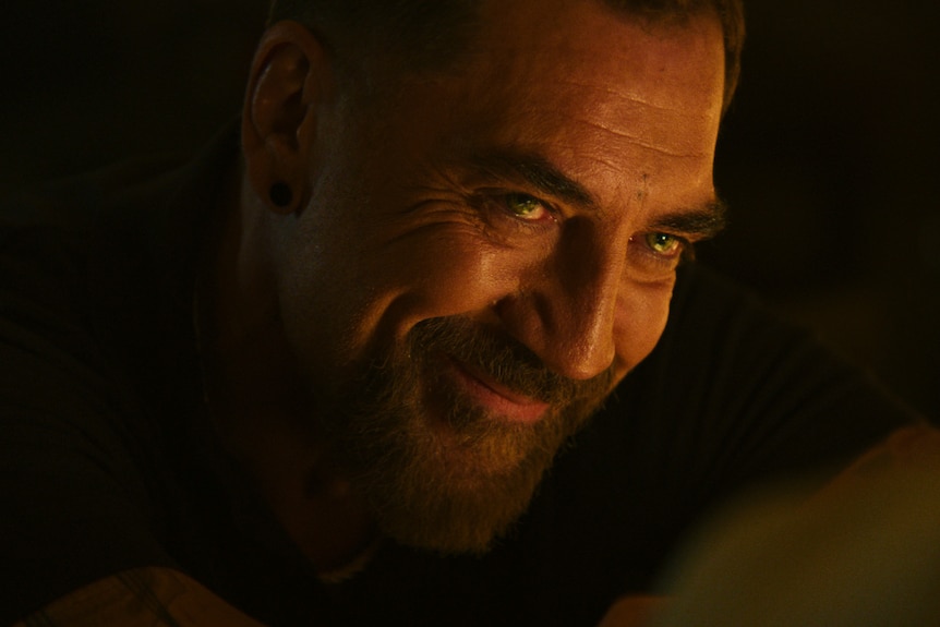 A TV still of Spanish actor Javier Bardem, 57, bearded, leans forward in dim light, with a sinister smile on his face.