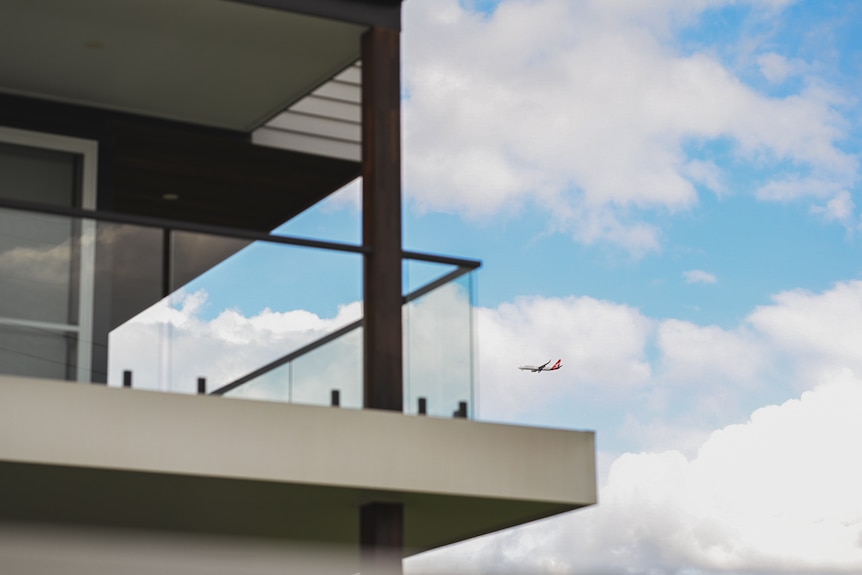 A plane flies through a cloudy sky, the deck of an apartment in the foreground.
