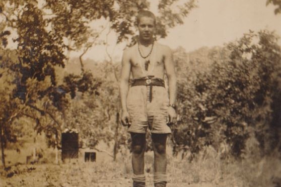Sepia image of young man in army boots, shorts, no shirt and crewcut standing above a trench and surrounded by trees