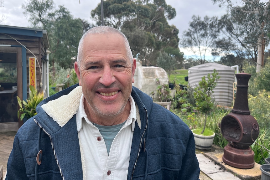 A man in white shirt with a blue fleece lined jacket in front of a garden.