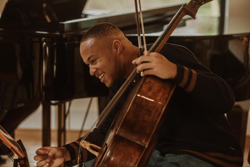 Sheku Kanneh-Mason sits holding a gleaming cello, one hand reaching towards a music stand. There's a grand piano in the room.