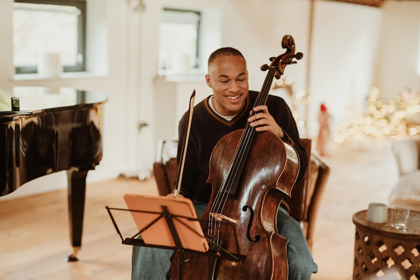 Sheku Kanneh-Mason sits holding a cello between his knees. One hand rests on the fingerboard, the other holds a bow.