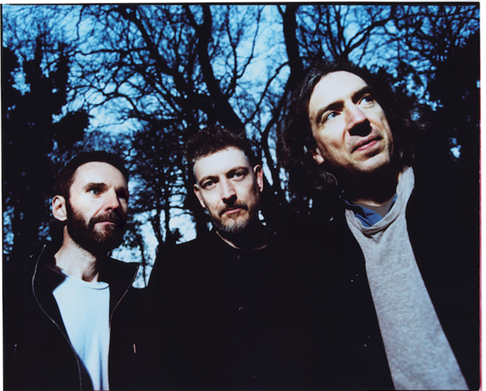 Promotional photo of Snow Patrol. The three members are standing against silhouetted tress, wearing a mixture of black and grey. 