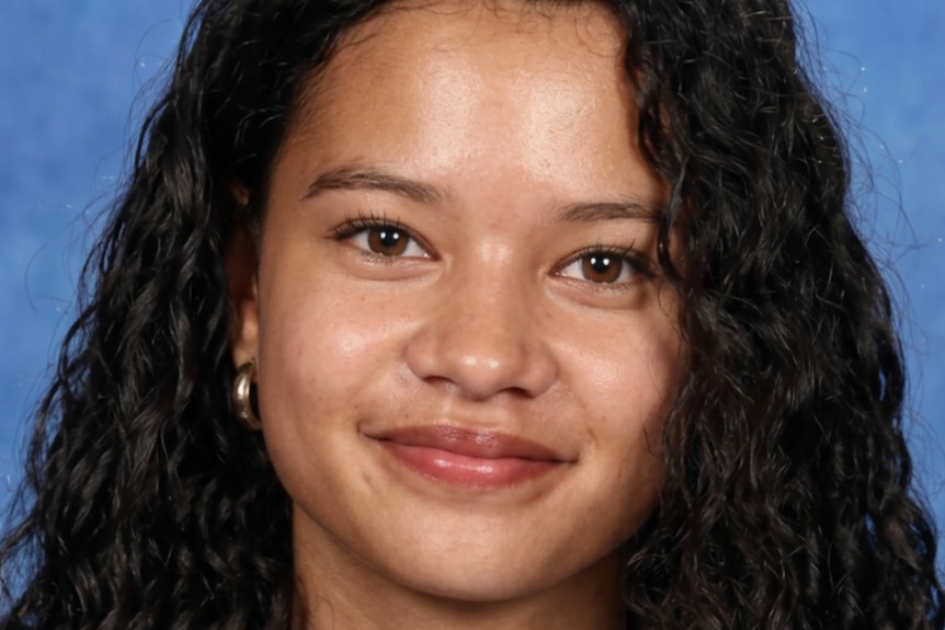 Laga'aia smiling while wearing gold hoop earrings and a white school shirt with long slightly curly hair.