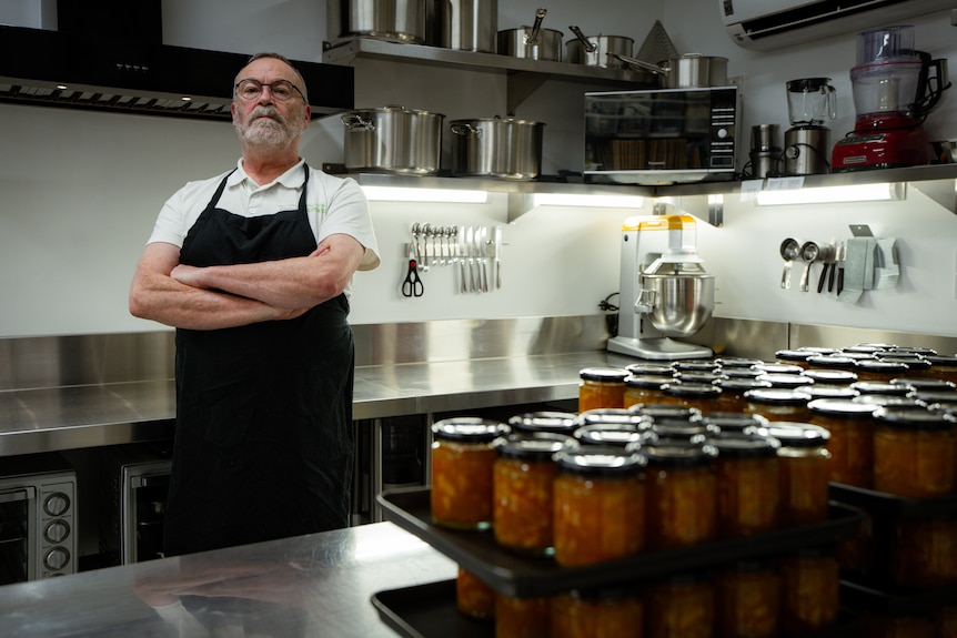 A bearded, middle-aged man in an apron stands near jars of produce in a commercial kitchen.
