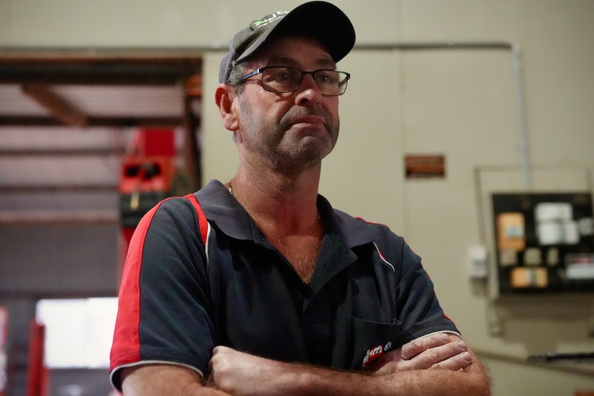 Middle aged man wearing black and red mechanic work shirt, black cap and glasses looks to right of frame in mechanic workshop