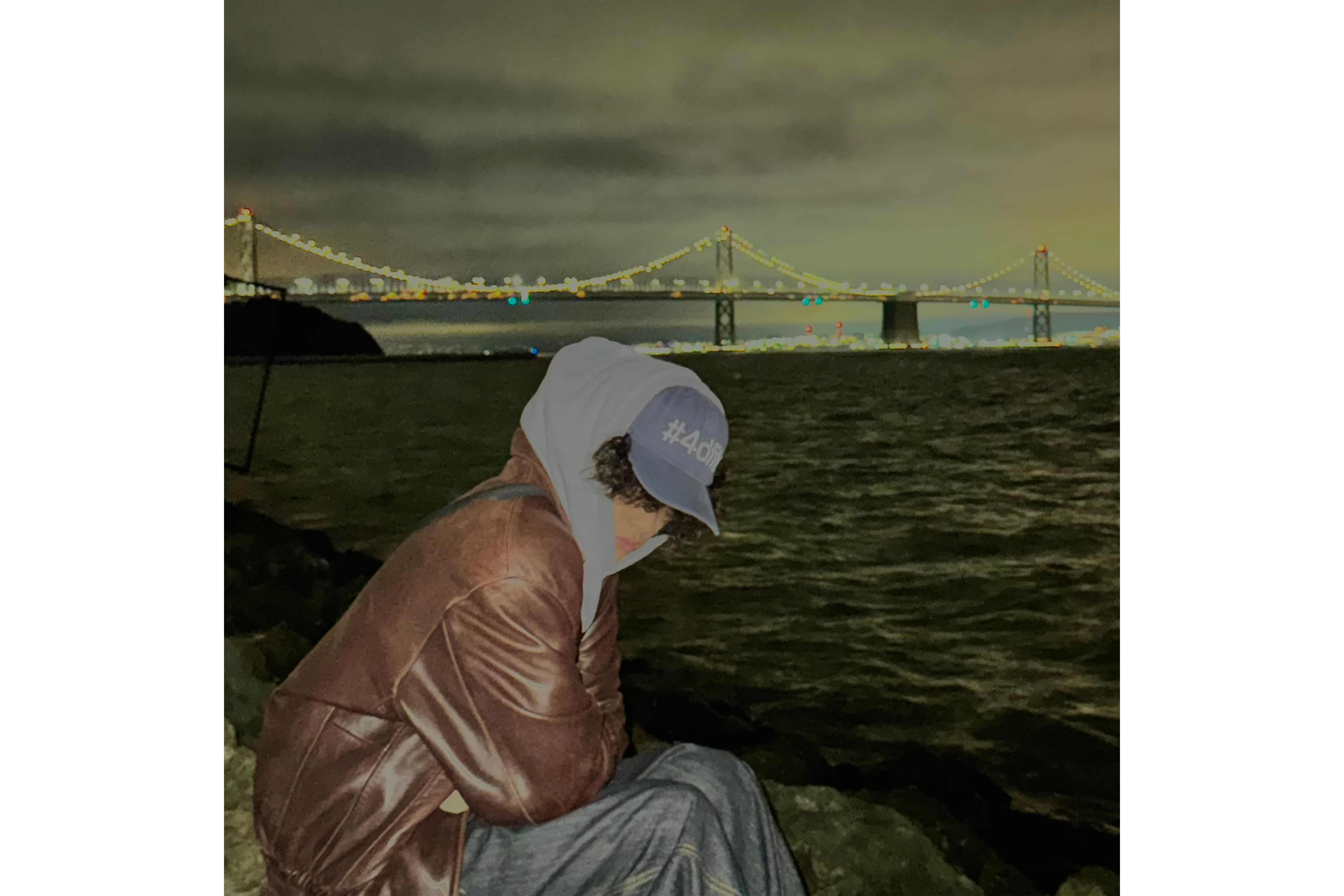 Dea sits on a rock in front of a waterfront with a bridge in the distance behind him. The singer said his Oakland upbringing influenced him to start by making hip-hop beats. (Courtesy of Dea/Altered from original)