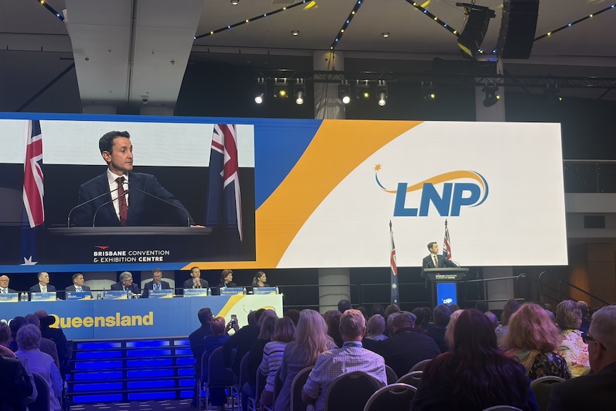 Premier David Crisafulli giving a speech at a lecturn with a large screen behind him.