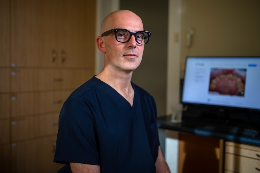 A man wearing blue scrubs and black glasses sits in front of a computer screen showing teeth.