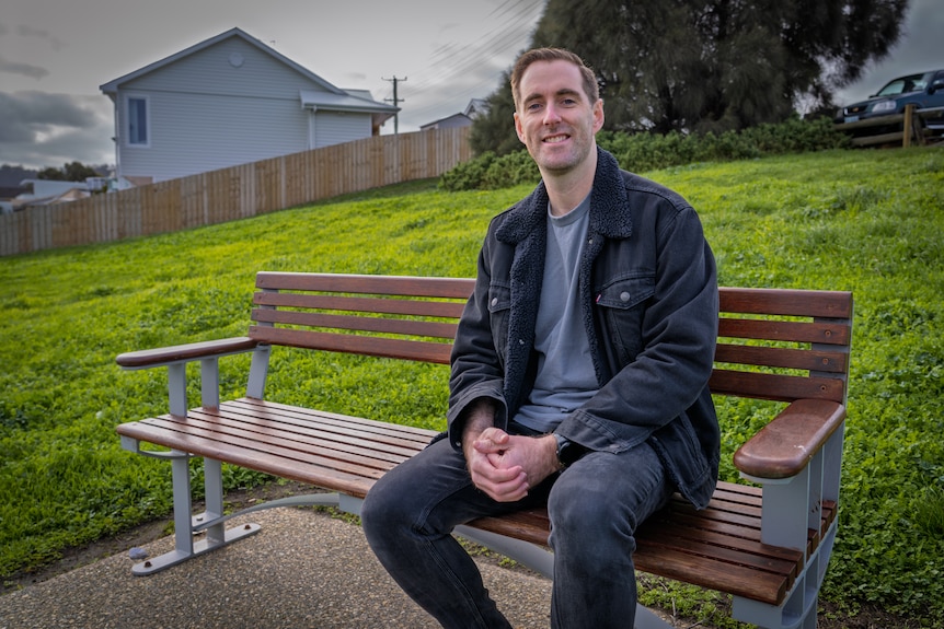 Jason Graham sits on a park bench and smiles at the camera.