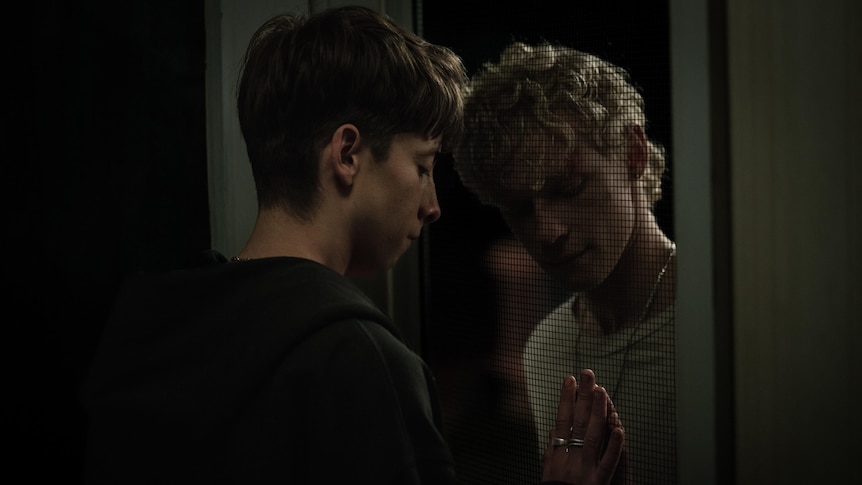 Two people lean their heads and hands together with a glass window between them