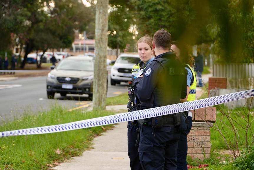 Three police officers stand behind police tape on the front verge of a house in a suburban street.