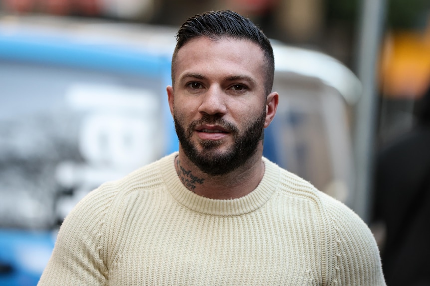 A dark-haired, bearded man in a knitted jumper walks down a city street.