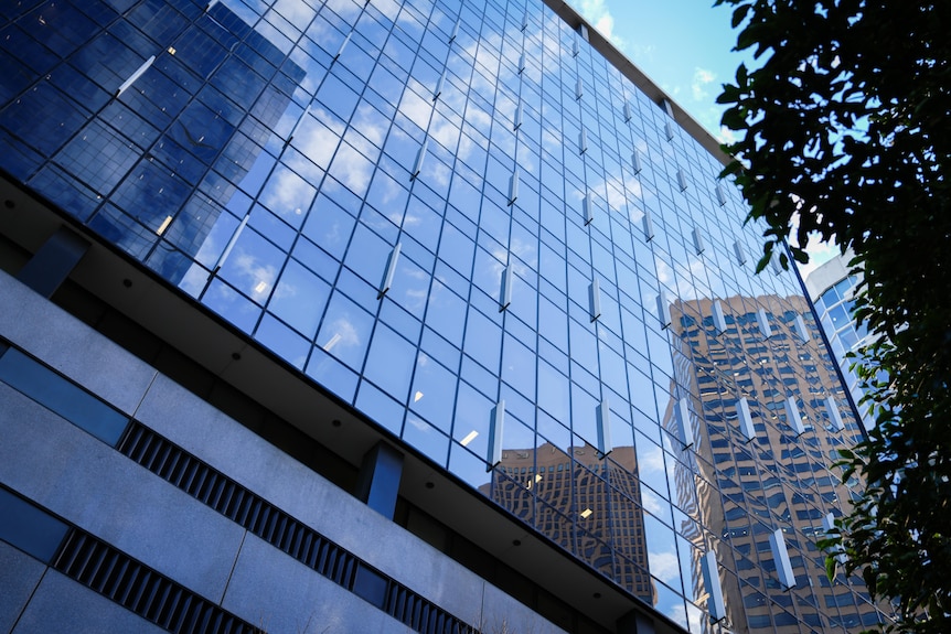 A picture taken looking up the glass facade of a multi-storey building that reflects highrises around it under a blue sky.