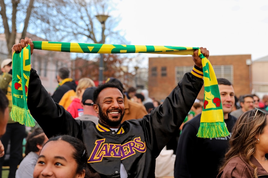 A man stands outside in a crowd of people, he is holding a green and gold scarf with Australia written on it above his head.