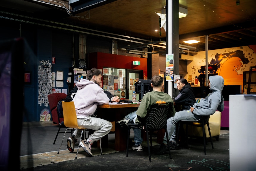 A group of 6 young people sit around a brown table in an open youth space with coloured walls, they're dressed casually.