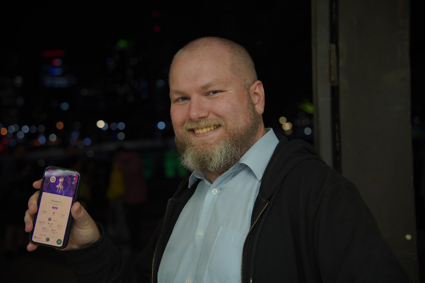 A bald man with a beard shows his phone screen against a night time backdrop