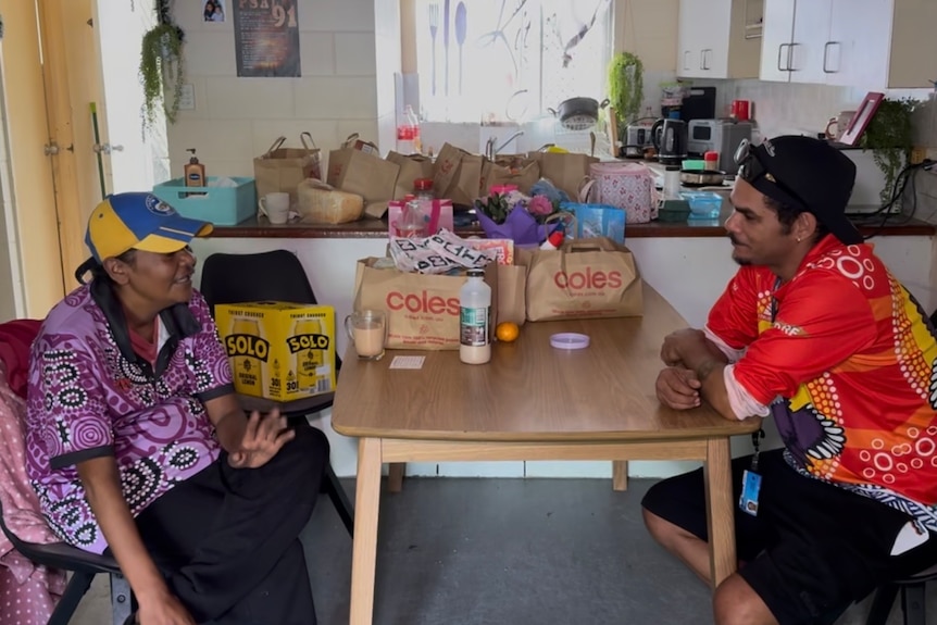 A woman and a man sit at a table with bags of groceries on the kitchen bench behind.