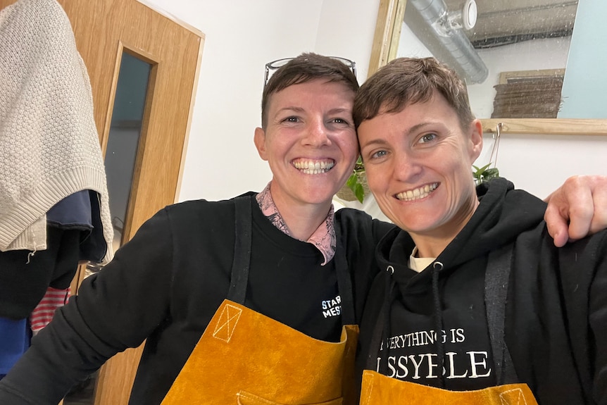 A female same-sex couple wearing black jumpers and aprons smile at the camera. The two women look very similar.