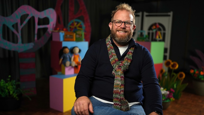 A man with a beard wears a sweater and scarf, seated and smiling. The colourful Play School set sits behind him.