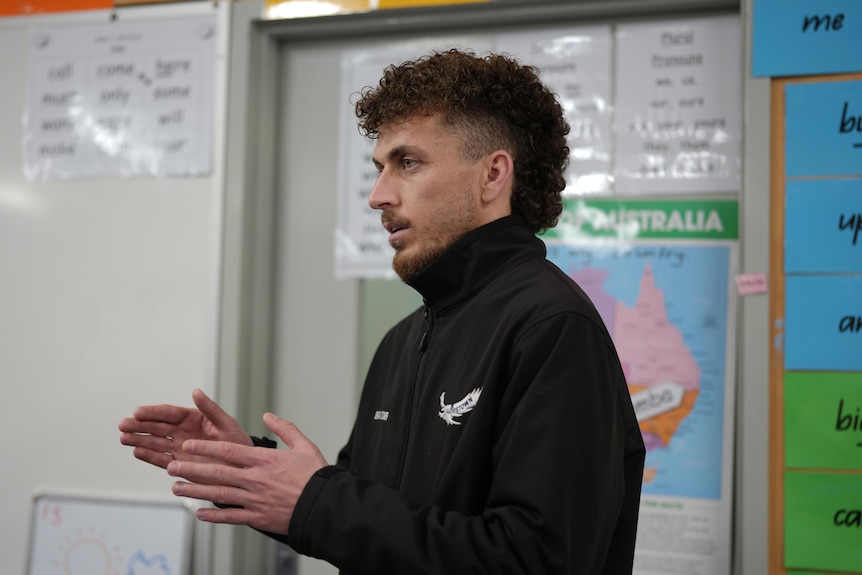 A man with curly hair wearing a black jacket speaking in a classroom.
