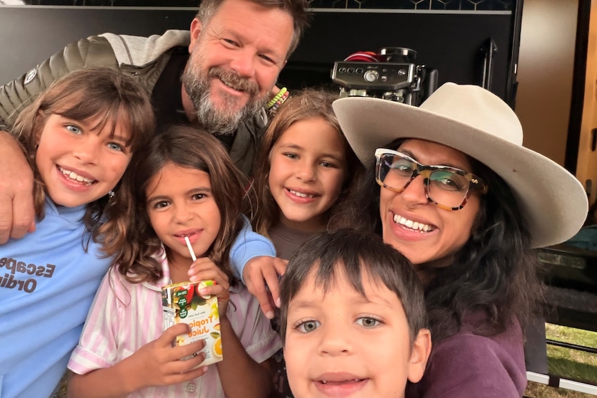 Tom and Huma Busby and their three daughters and one son stand in front of a caravan, smiling.