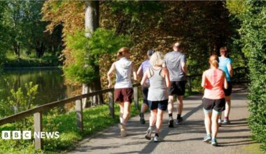 A group of people going for a run in parkland. There is a body of water to the left of the image, with trees and a small wooden fence