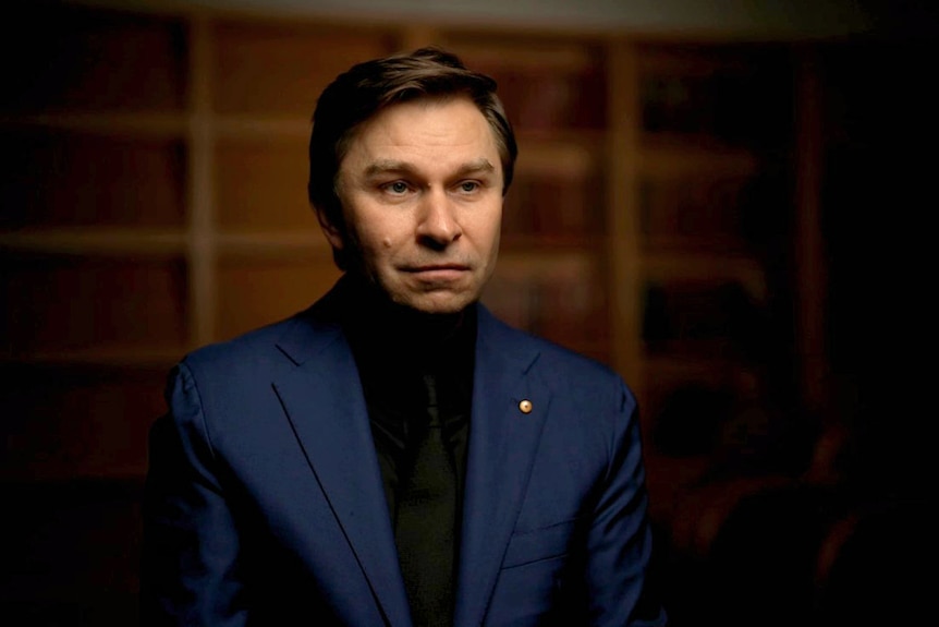David, is a man wearing a black shirt under a navy suit jacket, slight smile, well groomed.