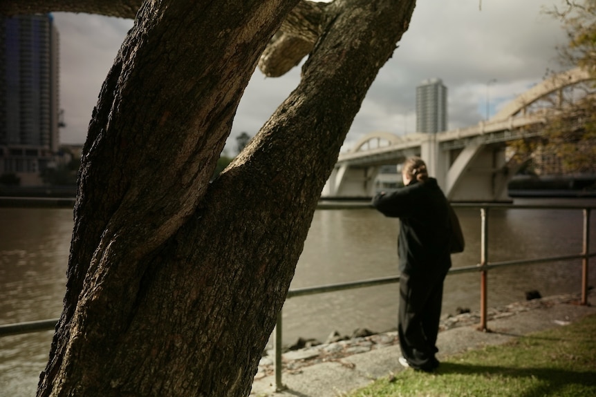 A woman stands on the edge of the Brisbane River leaning on a railing with a tree in focus in the foreground.