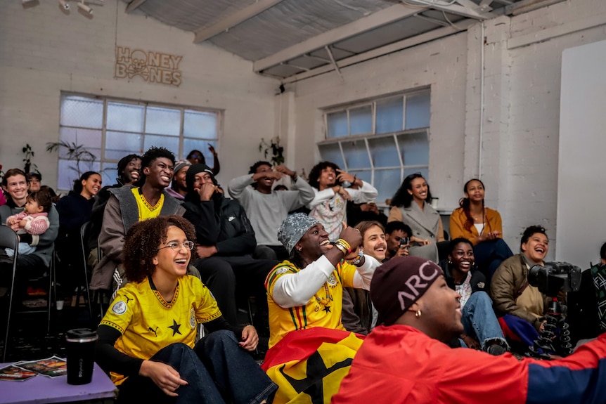 A group of people in a warehouse building look forward and cheer, some are wearing yellow shirts.